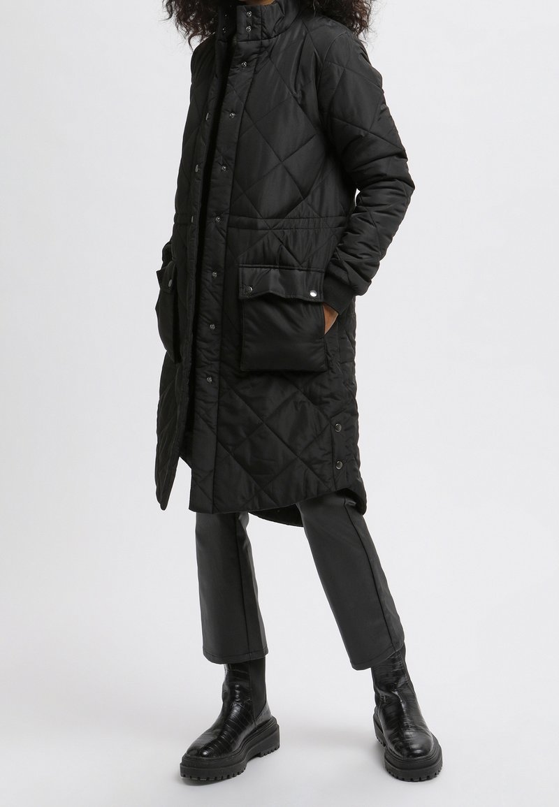 Abrigo largo negro acolchado con cuello alto, botones de presión, bolsillos laterales y un acabado elegante y brillante, combinado con pantalones negros ajustados y botas.