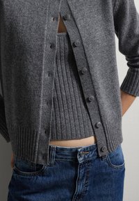Cardigan grigio realizzato in morbido knit, con orlo a coste e cinque bottoni scuri. Indossato sopra un top grigio a coste coordinato e jeans blu.