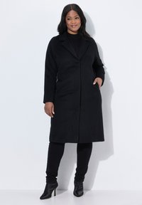 Cappotto di lana nero con rever a punta, tasche frontali e silhouette dritta. Indossato sopra un top e pantaloni neri, abbinato a stivali alla caviglia.