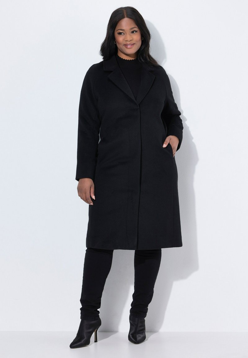 Cappotto di lana nero con rever a punta, tasche frontali e silhouette dritta. Indossato sopra un top e pantaloni neri, abbinato a stivali alla caviglia.