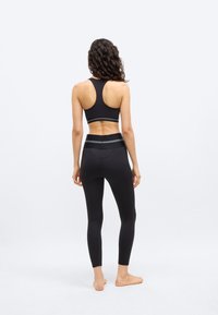 Donna in piedi a piedi nudi rivolta di spalle, indossa un reggiseno sportivo nero e leggings a vita alta coordinati con strisce grigie.