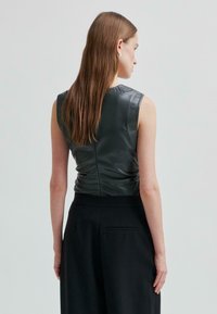 Žena s rovnými hnědými vlasy, oblečená v topu bez rukávů z černé kůže a černých kalhotách s vysokým pasem, otočená zády k jednobarevnému pozadí.