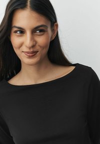 Fekete, hosszú ujjú felső, széles nyakkivágással, sima textúrával és minimalista designnal. A haj sötét és egyenes, enyhe hullámmal.