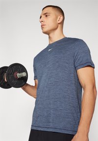 Meeste sinine spordipaita, millel on heatherdatud tekstuur, ümar kaelus, lühikesed varrukad ja väike logo rinnal, hoides hantlit.