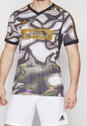 Mann trägt ein schwarzes, weißes und goldenes Adidas-Sporttrikot mit abstraktem Muster und dem Schriftzug „Love Unites“, kombiniert mit weißen Adidas-Shorts.