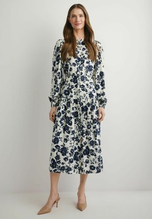 Kobieta o długich włosach, ubrana w białą sukienkę midi w czarne kwiatowe wzory oraz beżowe buty na wysokim obcasie, stojąca na jednolitym tle.