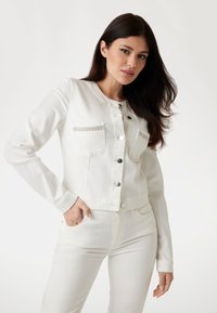 White denim jacket with silver button accents, two chest pockets with decorative trim, fit cropped at the waist, and long sleeves.