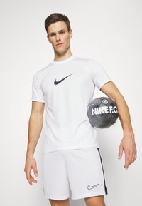 Vit Nike sporttröja med svart logotyp, kombinerad med matchande shorts. Modellen håller en grå Nike F.C. fotboll under armen. Slät tygtextur.