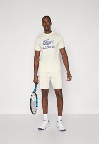 T-shirt a maniche corte color beige chiaro con logo Lacoste blu, abbinato a pantaloni corti beige e scarpe da ginnastica bianche, mentre tiene una racchetta da tennis.