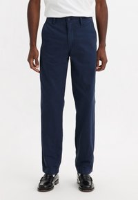 Pantalons en coton bleu marine avec une coupe droite, présentant un devant plat, des passants de ceinture et des poches latérales. Portés avec des mocassins noirs.