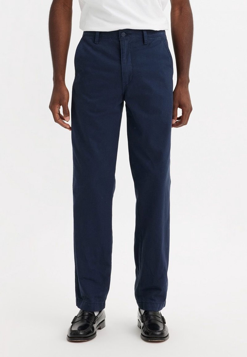 Pantalons en coton bleu marine avec une coupe droite, présentant un devant plat, des passants de ceinture et des poches latérales. Portés avec des mocassins noirs.