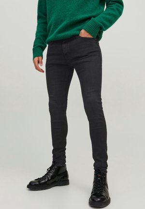 Jeans negros ajustados de tejido suave, con cinco bolsillos y pasadores para cinturón, combinados con botas negras de cordones y un suéter de punto verde.