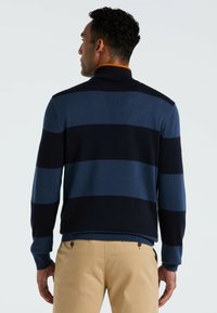 Man met zijn rug naar de camera, gekleed in een marineblauw en blauwe gestreepte trui met een oranje kraagdetail en beige broek, tegen een effen achtergrond.