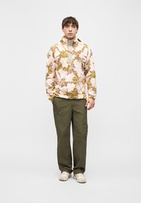 Νέος άνδρας φοράει ροζ και κίτρινη φλις μπλούζα με σχέδιο, χακί παντελόνι cargo και μπεζ αθλητικά, στέκεται πάνω σε λευκό φόντο.