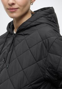 Μαύρο καπιτονέ μπουφάν με κουκούλα. Διαθέτει ραφές σε σχήμα διαμαντιού, λεπτομέρεια με κορδόνι και κλείσιμο με κουμπιά πιέσεως. Λεία υφή.