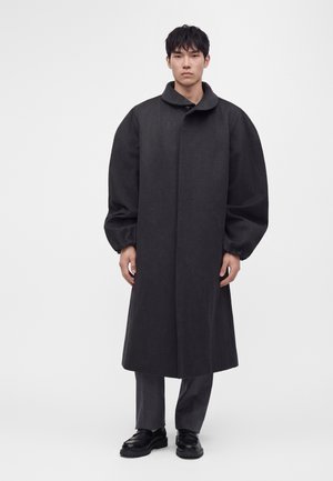 Jeune homme debout portant un manteau gris foncé surdimensionné à larges manches par-dessus un pantalon gris et des chaussures noires sur un fond uni.