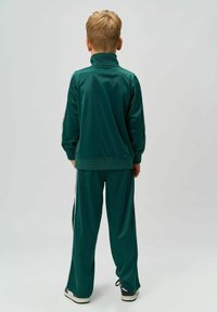 Completo sportivo verde con alta collo e polsini a costine; presenta bande laterali ed è abbinato a sneakers nere.
