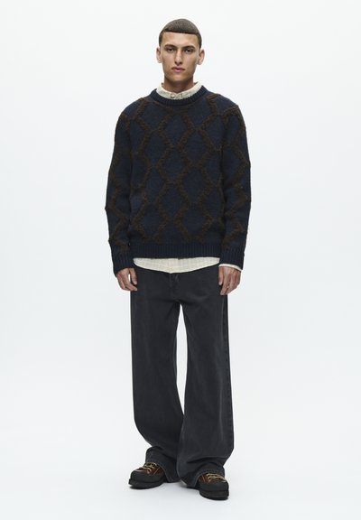 Granatowy sweter dziany z brązowym wzorem w romby, zestawiony z luźnymi ciemnoszarymi dżinsami i masywnymi czarnymi butami. Nałożony na białą koszulę.