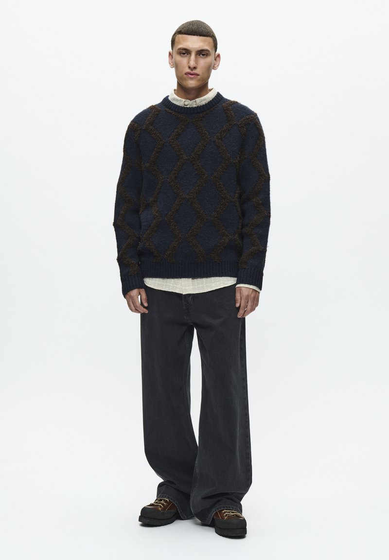 Pull en maille bleu marine avec motif losange marron, assorti à un jean gris foncé ample et des chaussures noires épaisses. Superposé à une chemise blanche.