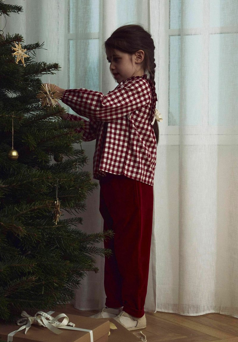 Un niño con una camisa de cuadros rojos y pantalones de terciopelo rojos decora un árbol de Navidad con adornos dorados, junto a regalos y cortinas transparentes.
