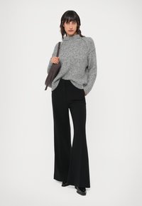 Suéter gris de punto con cuello alto, combinado con pantalones de campana negros. La modelo sostiene un bolso gris oscuro, de pie frente a un fondo blanco.