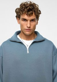 Lichtblauwe ritstrui met hoge kraag, gemaakt van een gestructureerde stof, met verlaagde schouders en een zilveren rits. Gelaagd over een wit shirt.