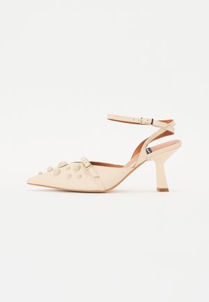 Beige pointed-toe slingback shoe with a textured upper featuring round studs, adjustable ankle strap, and a thick, tapered heel.
