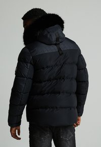 Mørkeblå dunjakke med faux fur hette, quiltet tekstur og justerbar stropp på baksiden. Har en logo på ermet.