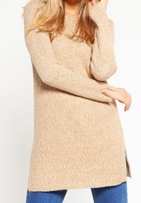 Pull en tricot beige avec une texture finie, à manches longues et fentes sur les côtés. Tissu épais et doux, offrant une coupe décontractée et un look casual.