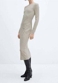 Nainen, jolla on päällään tiukka pitkähihainen beige mekko mustilla vaakasuorilla raidoilla ja mustat polvipituiset korkeat saappaat, seisoo valkoista taustaa vasten.