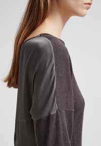 Vêtement gris foncé avec une texture douce, présentant des panneaux d'épaule gris lisse contrastants. Design simple avec une coupe décontractée.