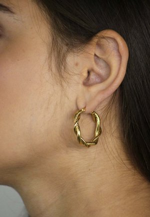 Goldener Creolen-Ohrring mit gedrehtem Design und strukturierter Oberfläche, getragen am Ohr eines Modells, der seine runde Form und glänzende Oberfläche hervorhebt.