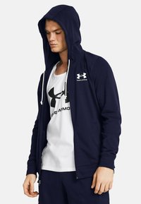 Felpa con zip navy e coulisse, con logo Under Armour; tessuto soffice, vestibilità regolare, abbinata a una canotta bianca con logo nero.