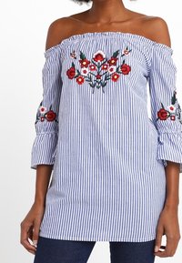 Sininen ja valkoinen raidallinen olkaimeton paita, jossa on kukkiteemaista brodeerausta hihoissa, rypytetty hihansuuhun ja rento istuvuus.