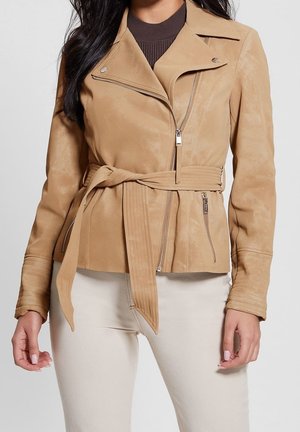 Femme portant une veste marron clair ceinturée avec des poches zippées sur un haut sombre et un pantalon blanc, vue des épaules aux hanches.