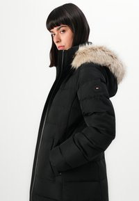 Cappotto imbottito nero con cappuccio foderato in pelliccia sintetica, chiusura con zip e tasche laterali. Presenta un motivo trapuntato e un accento con logo sulla manica sinistra.