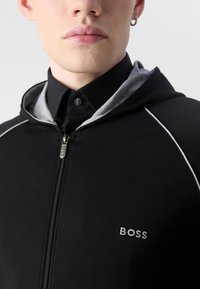 Svart hettegenser med grå detaljer, med en logo på brystet, lett materiale og design med dobbel hette.