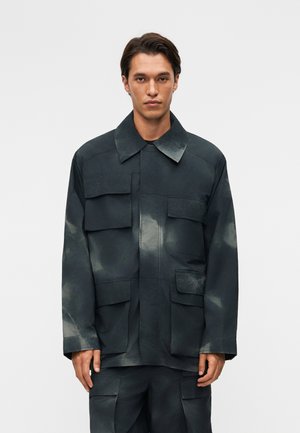 Νέος άνδρας φοράει μαύρο υπερμεγέθη μπουφάν και ταιριαστό παντελόνι με πολλές μεγάλες τσέπες με καπάκια, στέκεται απέναντι σε απλό γκρι φόντο.