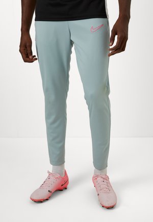 Light blue athletic pants with pink Nike logo, tapered fit, elastic waistband, and ribbed ankle cuffs. Worn with pink soccer cleats.