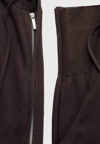 Hoodie marrone scuro con zip argento, caratterizzato da polsini a coste e tessuto testurizzato. Inquadratura ravvicinata della zip e del bordo vita.