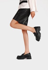 Scarpe di pelle nera con suola spessa, design senza lacci e una fascia decorativa. Il modello indossa una gonna in pelle nera e una maglietta bianca.