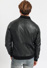 Man med ljusbrunt hår som bär en svart läderjacka och bleknade blå jeans, vänd bort från kameran mot en enkel bakgrund.