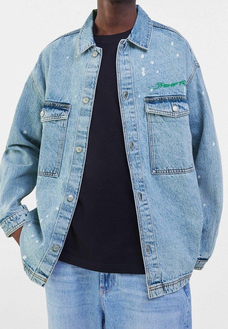 Ljusblå jeansjacka med avslappnad passform, med detaljer av färgstänk och grön brodyr på bröstet, knäppt fram och två fickor.