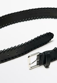 Zwart gevlochten leren riem met een zilverkleurige metalen gesp. De riem heeft een gestructureerd oppervlak met gelijkmatig verdeelde gaatjes voor aanpassing.