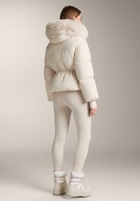 Nafúkaná krémová bunda s kapucňou lemovanou kožušinou, elastickým pásom a rebrovanými lemami, doplnená priliehavými krémovými legínami a bielymi zimnými topánkami.