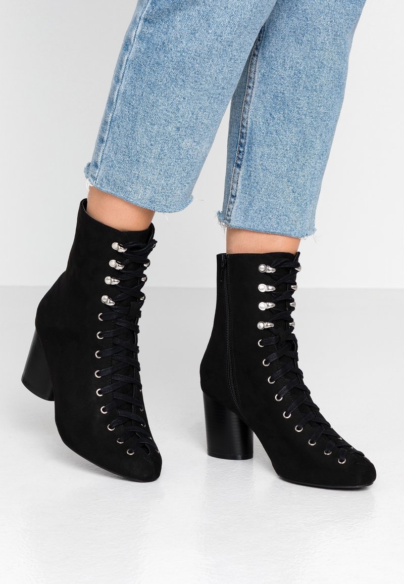 Botas de ante negras hasta el tobillo con puntera afilada, frente con cordones, ojales plateados y tacón grueso. Combinadas con unos vaqueros cortos de mezclilla azul claro.
