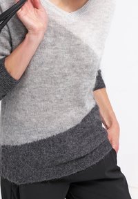 Persona che indossa un maglione in maglia morbida a blocchi di colore in grigio chiaro e scuro, con una tracolla nera su una spalla e una mano nella tasca.