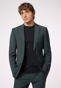 Completo su misura verde scuro con rever strutturati, chiusura a un bottone e camicia nera sotto. Presenta un sottile accento rosso sul rever.