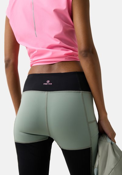 Kvinde iført pink top og tofarvede grønne og sorte træningsleggings, der holder en foldet jakke i den ene hånd.