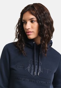 Felpa navy con logo ricamato, cappuccio con coulisse e polsini a coste. Tessuto morbido, vestibilità comoda e dettaglio con design a scudo sulla parte frontale.